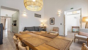 A spacious living area with a long wooden table, woven mats, a sofa, a chair, and a hanging light fixture.