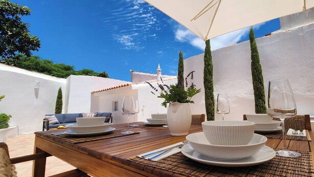 An outdoor dining area with a wooden table set for two, white plates, bowls, and wine glasses.