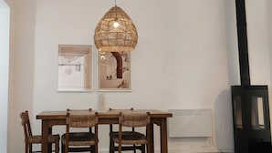 A dining area with a wooden table and chairs, a woven pendant light, framed artwork on the wall, and a black chimney.