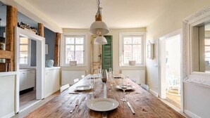 A dining area with a rustic wooden table set for four, flanked by windows with shutters, a hanging light fixture, and a view into another room. - Stilhaus 1730 - Central. Quiet. Unique. 1st floor (Königsbach-Stein)