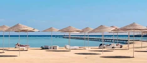 A beach with umbrellas and lounge chairs, a pier extending into the water, and a clear sky.
