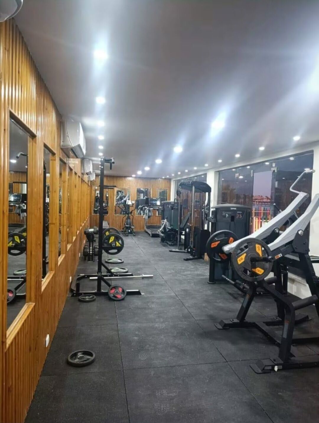 Et fitnessrum med forskellig træningsudstyr, træpaneler på den ene væg og store vinduer.