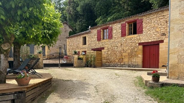 A stone building with red shutters, a gravel driveway, and a wooden deck with chairs. - Farm cottage (LALINDE)