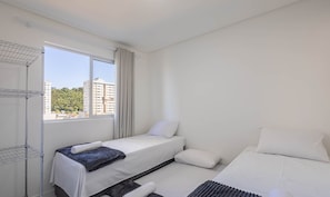 Habitación de hotel con cama, una mesita con una toalla, ventana con cortinas y vistas a edificios y vegetación.