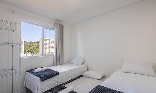 Ett hotellrum med en säng, ett litet bord med en handduk, ett fönster med gardiner och utsikt över byggnader och grönska utanför.