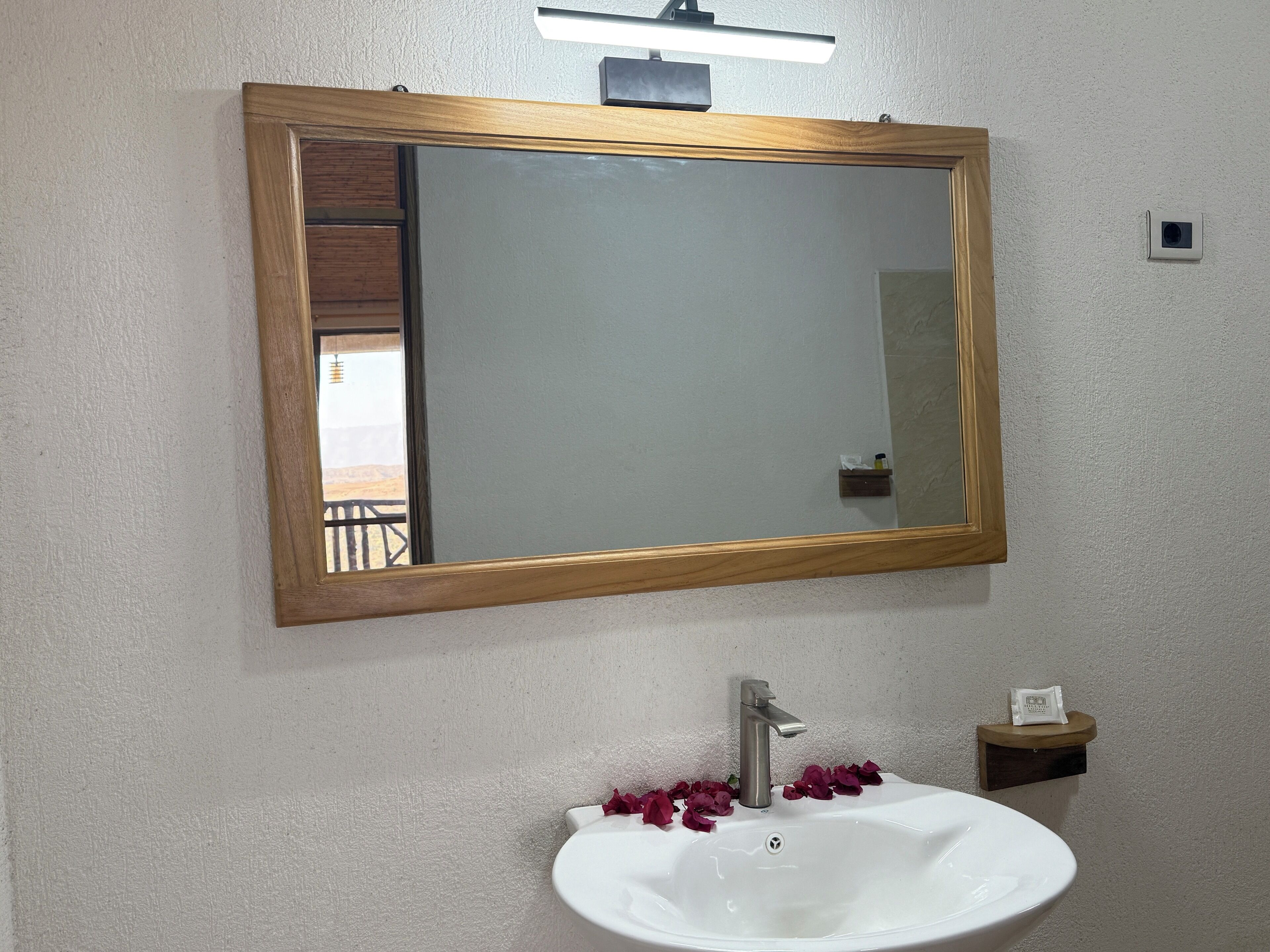 Un baño con lavamanos blanco, un espejo con marco de madera y un estante pequeño con artículos de aseo.