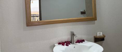 Un baño con lavamanos blanco, un espejo con marco de madera y un estante pequeño con artículos de aseo.