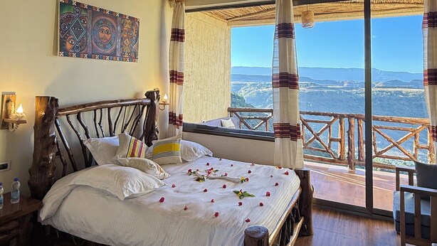 A rustic wooden room with a bed, a nightstand, a painting, and a balcony with a view. - Hilltop Lodge - Lalibela  (Lalibela)