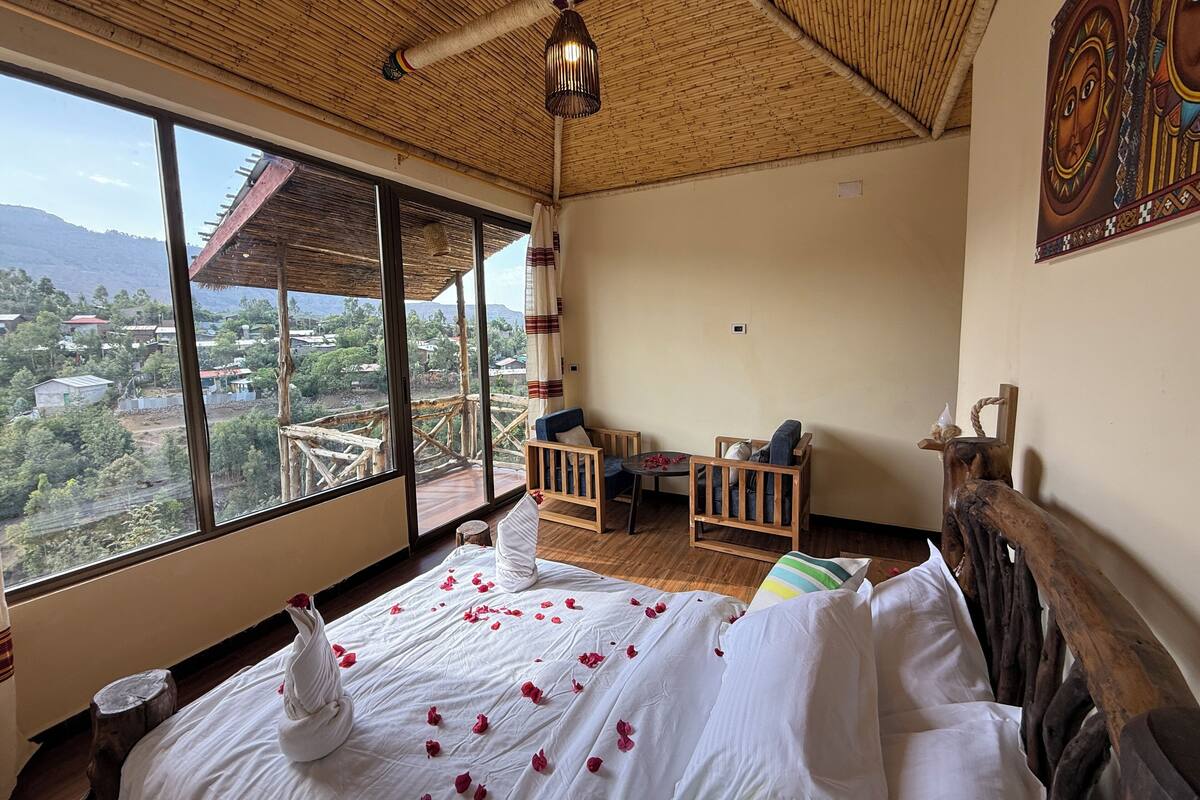 Una habitación con una cama decorada con pétalos de rosa, techo de madera, vistas a un pueblo a través de amplios ventanales y un mural tradicional en la pared.
