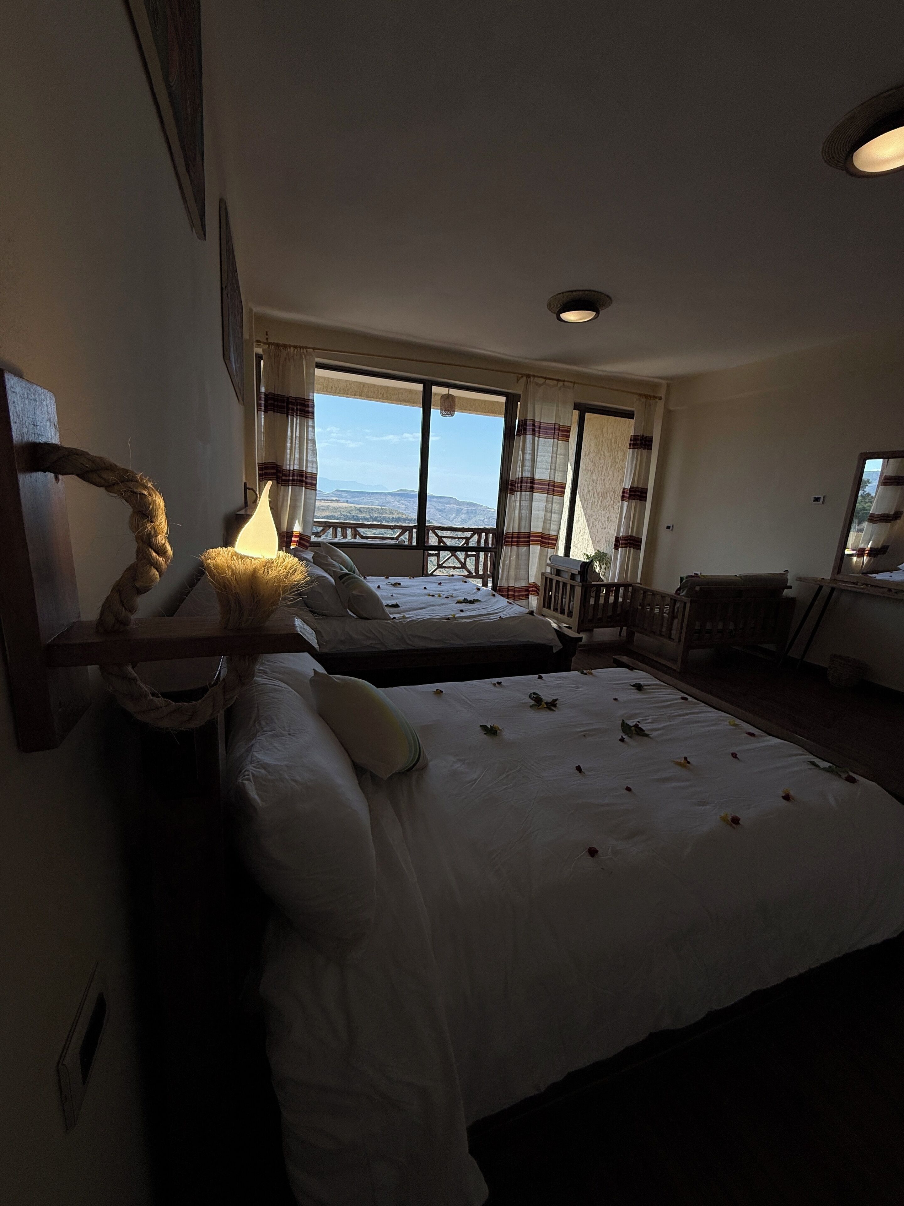 A hotel room with two beds, a large window with a view, a wooden headboard, and a bedside lamp.