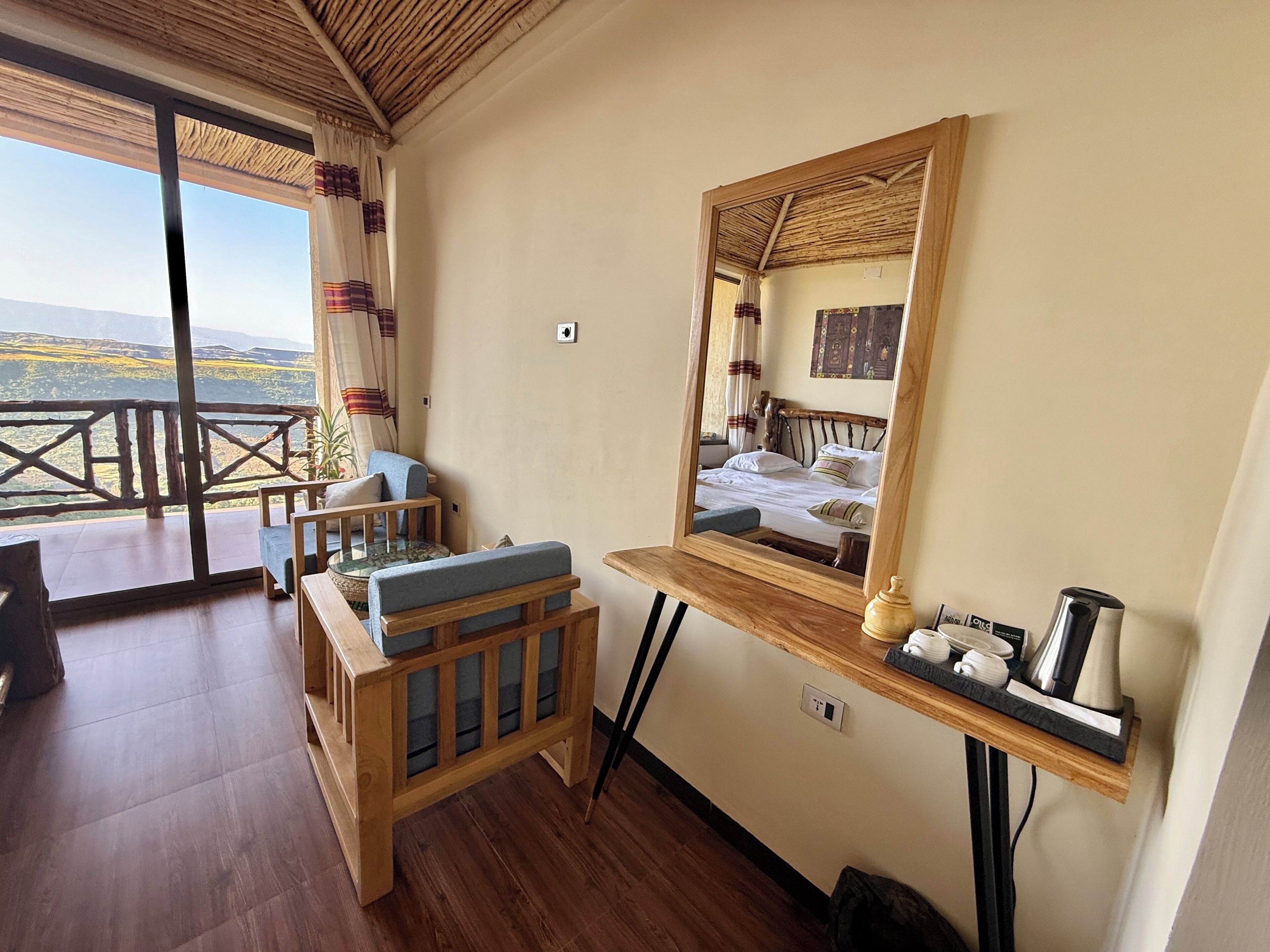 Una habitación con un escritorio de madera, una cama con cabecera, un espejo y un balcón con vistas al paisaje.