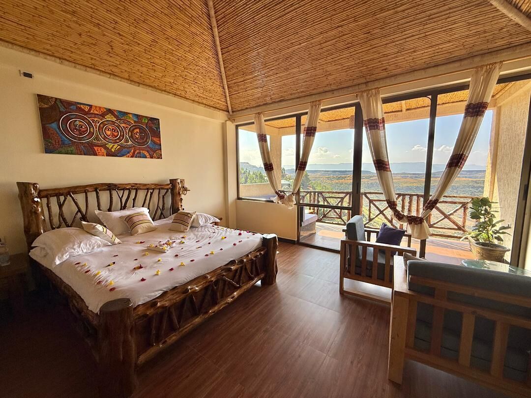 Une chambre avec un lit en bois, une vue dégagée sur l’extérieur grâce à de grandes fenêtres et un tableau accroché au mur.