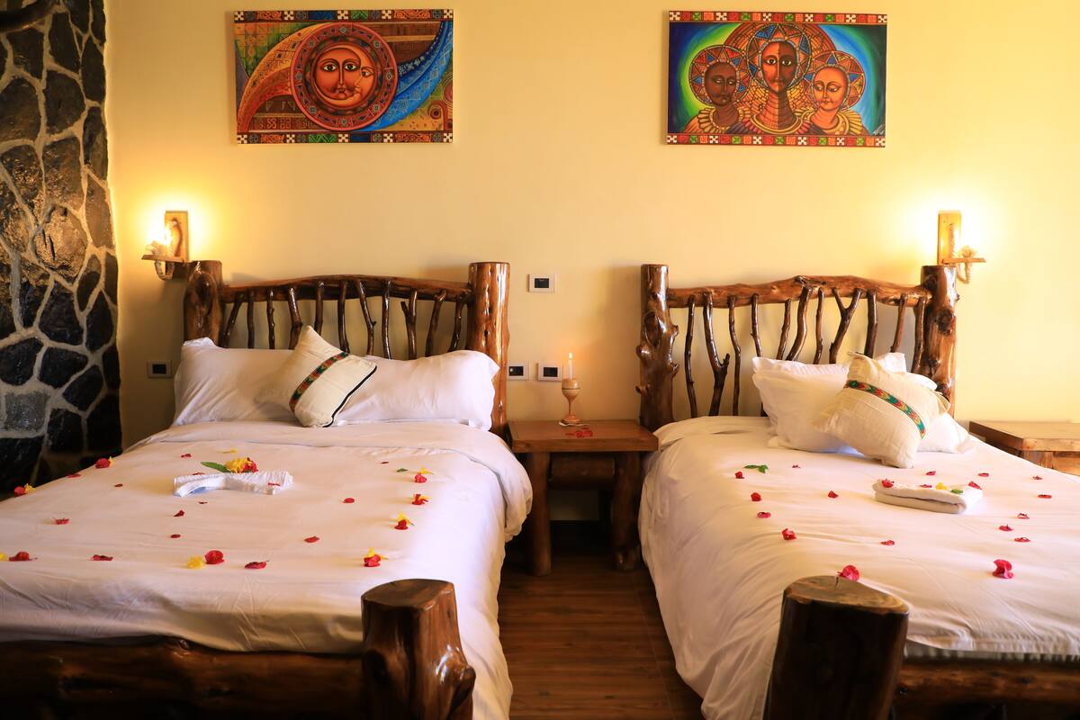 Habitación de hotel con dos camas de madera, ropa de cama blanca y almohadas decorativas. Dos cuadros adornan la pared sobre las camas.