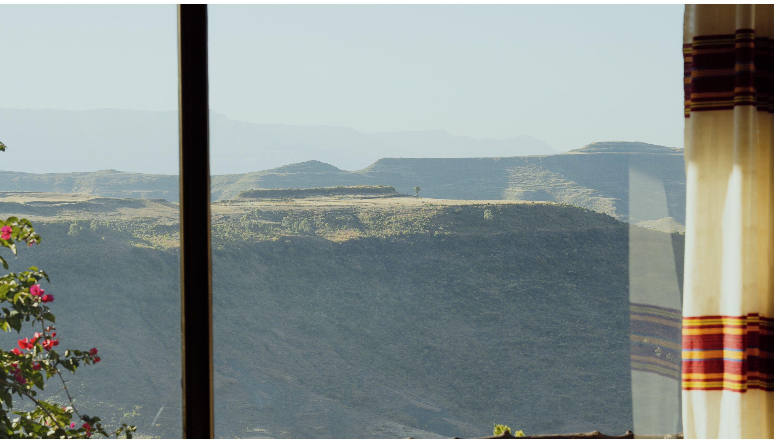 A view of a mountainous landscape through a window with a curtain partially visible on the right.