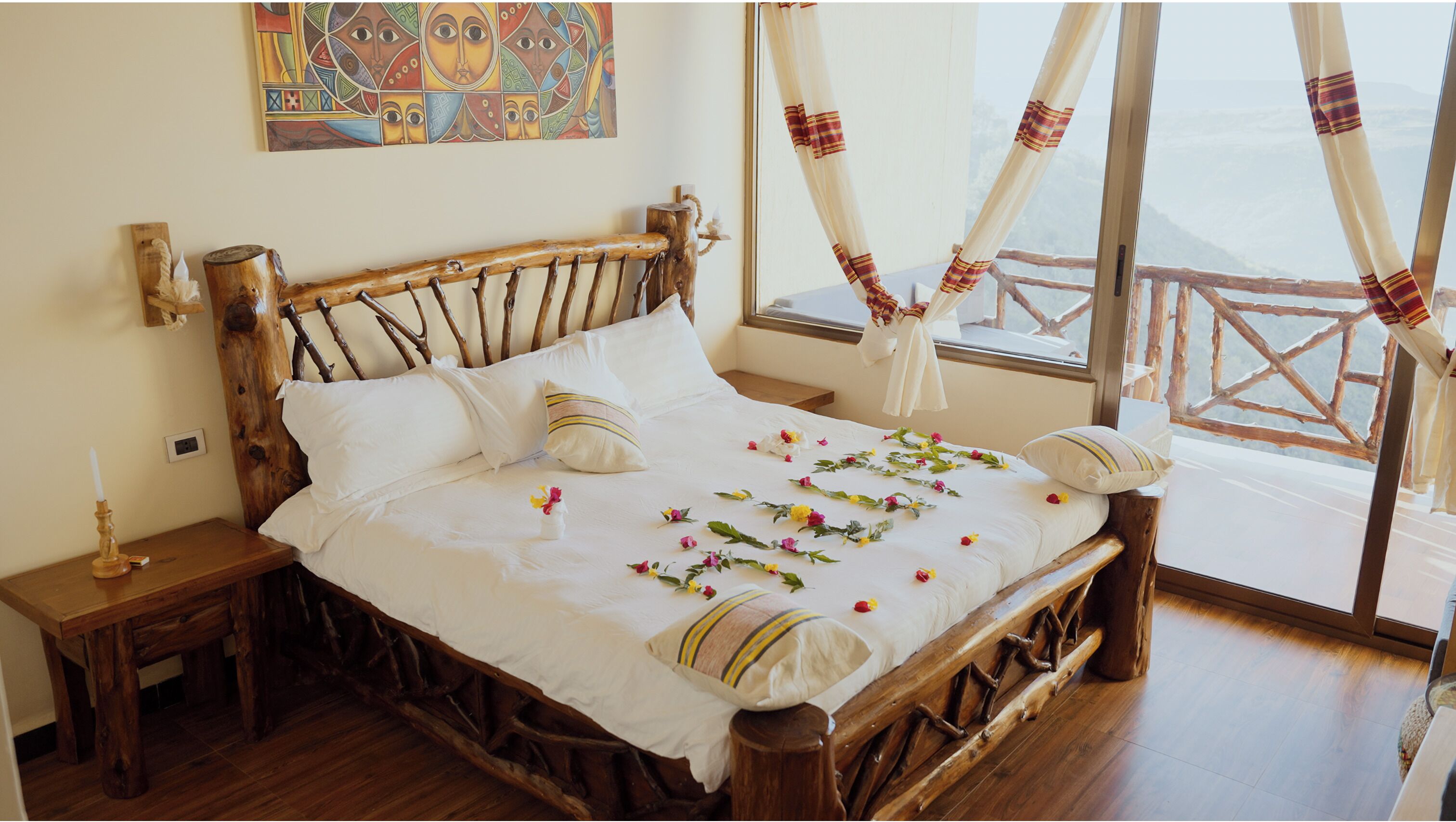 Una cama de madera con cubrecama y almohadas blancas, una mesita de noche con una vela y vistas al exterior a través de una ventana con cortinas.