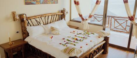 Una cama de madera con cubrecama y almohadas blancas, una mesita de noche con una vela y vistas al exterior a través de una ventana con cortinas.