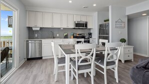 A modern kitchen with white cabinets, a stainless steel dishwasher, and a dining area with white chairs and a table. - Oceanfront home with breathtaking views, West! (Emerald Isle)