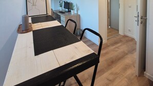A modern dining area with a wooden table, black chairs, and a black placemat. There is a clock on the wall and a small desk with a computer and plants.