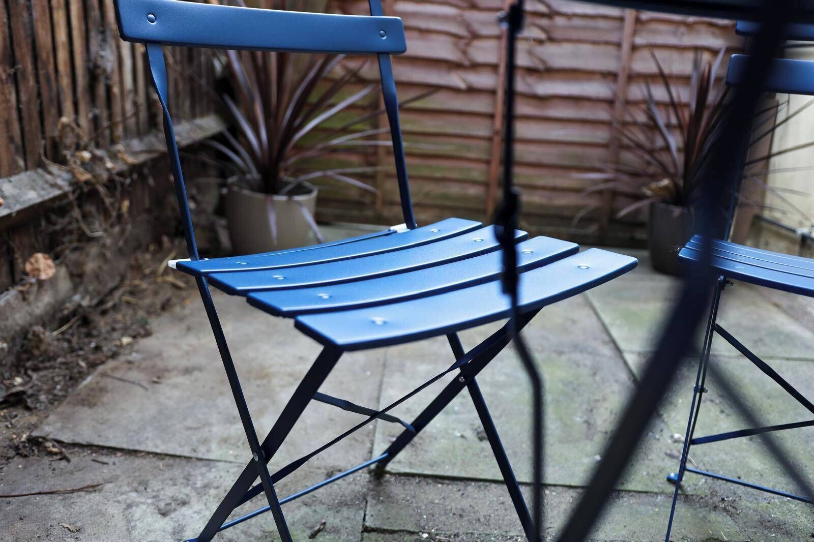 Una silla plegable de metal azul, con respaldo y reposabrazos, ubicada en un patio de concreto con una valla de madera y plantas en macetas al fondo.