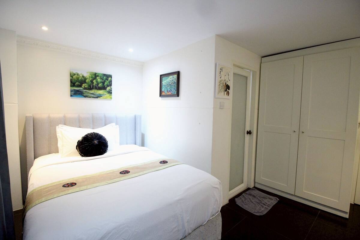 A neatly made bed with a white comforter and a decorative pillow, a small painting on the wall, and a closed wardrobe.