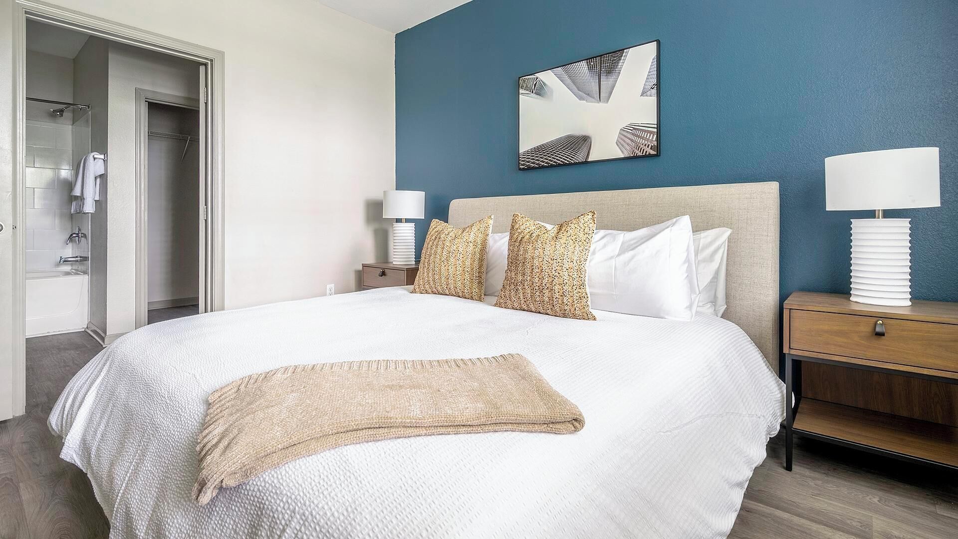 A hotel room with a large bed, a nightstand, a bedside table, a lamp, a painting on the wall, and a bathroom with a shower and a towel rack.