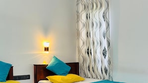 A hotel room with two wooden beds, each with a teal bedspread and pillows in yellow and blue. There is a wall-mounted light fixture on the left and a patterned curtain on the right. - Blue Sky Hotel Yala (Mahasenpura)