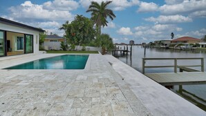 A swimming pool with a wooden deck and a dock extending into a body of water, surrounded by residential buildings and palm trees. - Parrots Cove (St. Pete Beach)