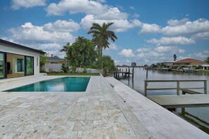 A swimming pool with a wooden deck and a dock extending into a body of water, surrounded by residential buildings and palm trees.