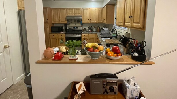 Une cuisine avec des meubles de rangement en bois, un four à micro-ondes, un four traditionnel et un plan de travail sur lequel on trouve des fruits et une radio.