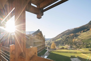 A wooden building with a sloped roof, a visible balcony, and a view of a green valley with houses and trees.