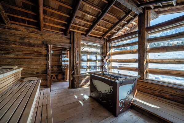 Interior de una cabaña de troncos con sauna, pisos de madera y grandes ventanales con vista a un paisaje nevado.