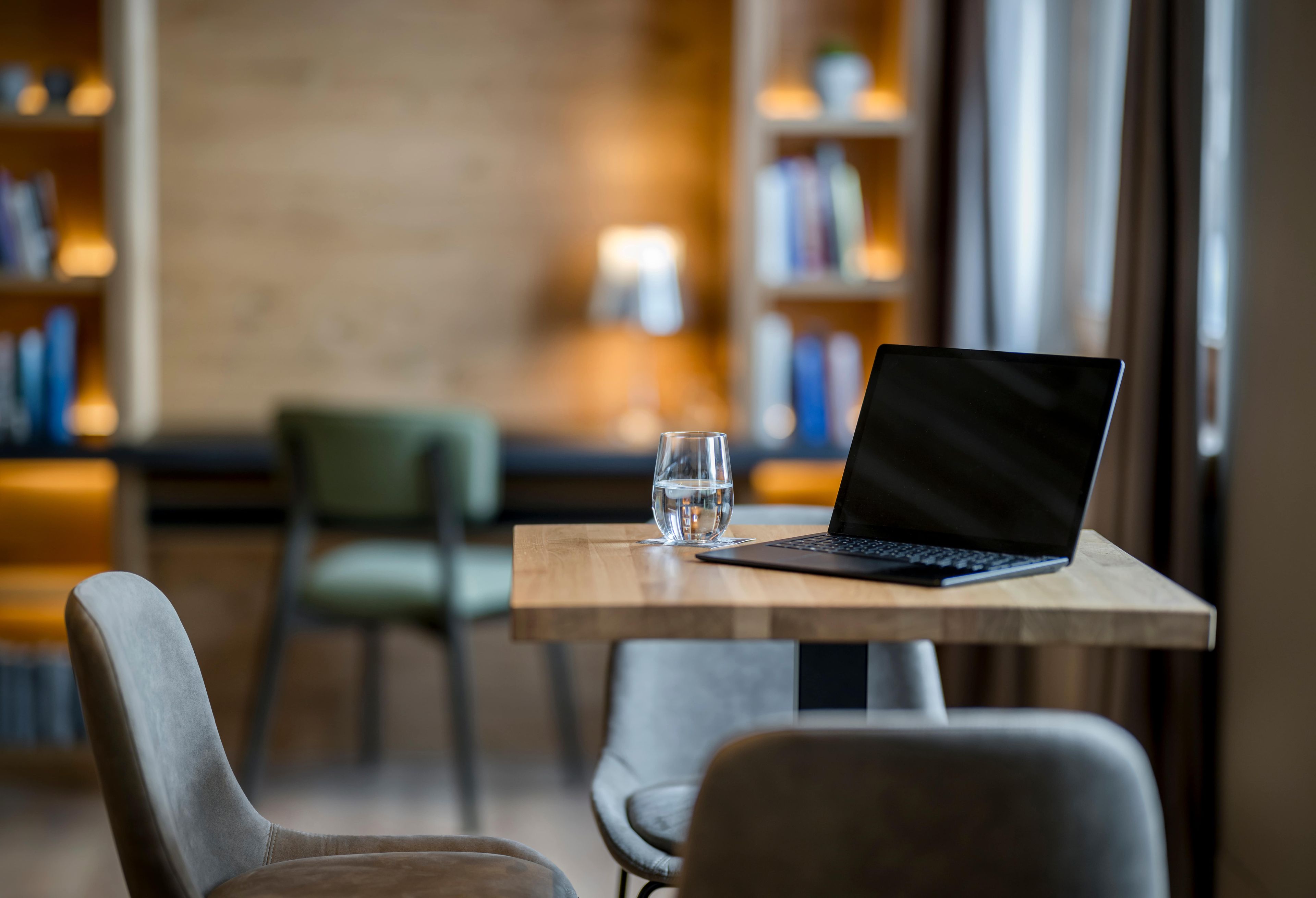 A laptop on a wooden table with a glass of water beside it, in a room with chairs and bookshelves in the background.