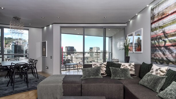 Un salon moderne avec un grand canapé, une table à manger et des chaises. Il y a un balcon avec vue sur la ville.
