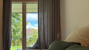 A room with a bed, a window with curtains, and a view of greenery and buildings outside. - The SeeQuartier - A place to arrive, take a deep breath and enjoy Lake Starnberg (Seeshaupt)