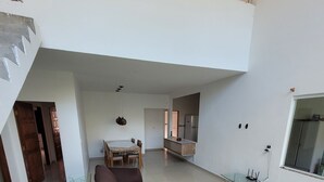 Un vaste espace de vie comprenant un canapé marron, une table basse en verre et une table à manger avec des chaises. Il y a également un coin cuisine avec un réfrigérateur et une fenêtre.