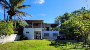 Une maison moderne à deux étages, dotée d’un porche abrité, entourée d’une végétation luxuriante et d’un palmier.