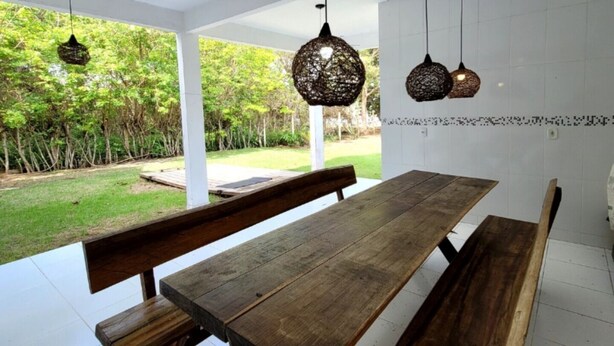 Une vaste terrasse dotée d’une table en bois et de bancs, entourée de murs blancs et agrémentée de luminaires suspendus.