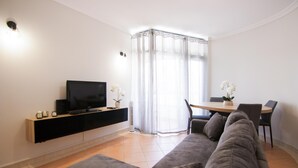 A modern living room with a flat-screen TV, a sofa, a dining table with chairs, and a large window with sheer curtains. - Modern 3-Bedroom Retreat in. Faro’s City Center (Faro)