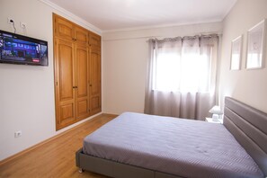 A bedroom with a bed, wooden wardrobe, television mounted on the wall, and a window with curtains.