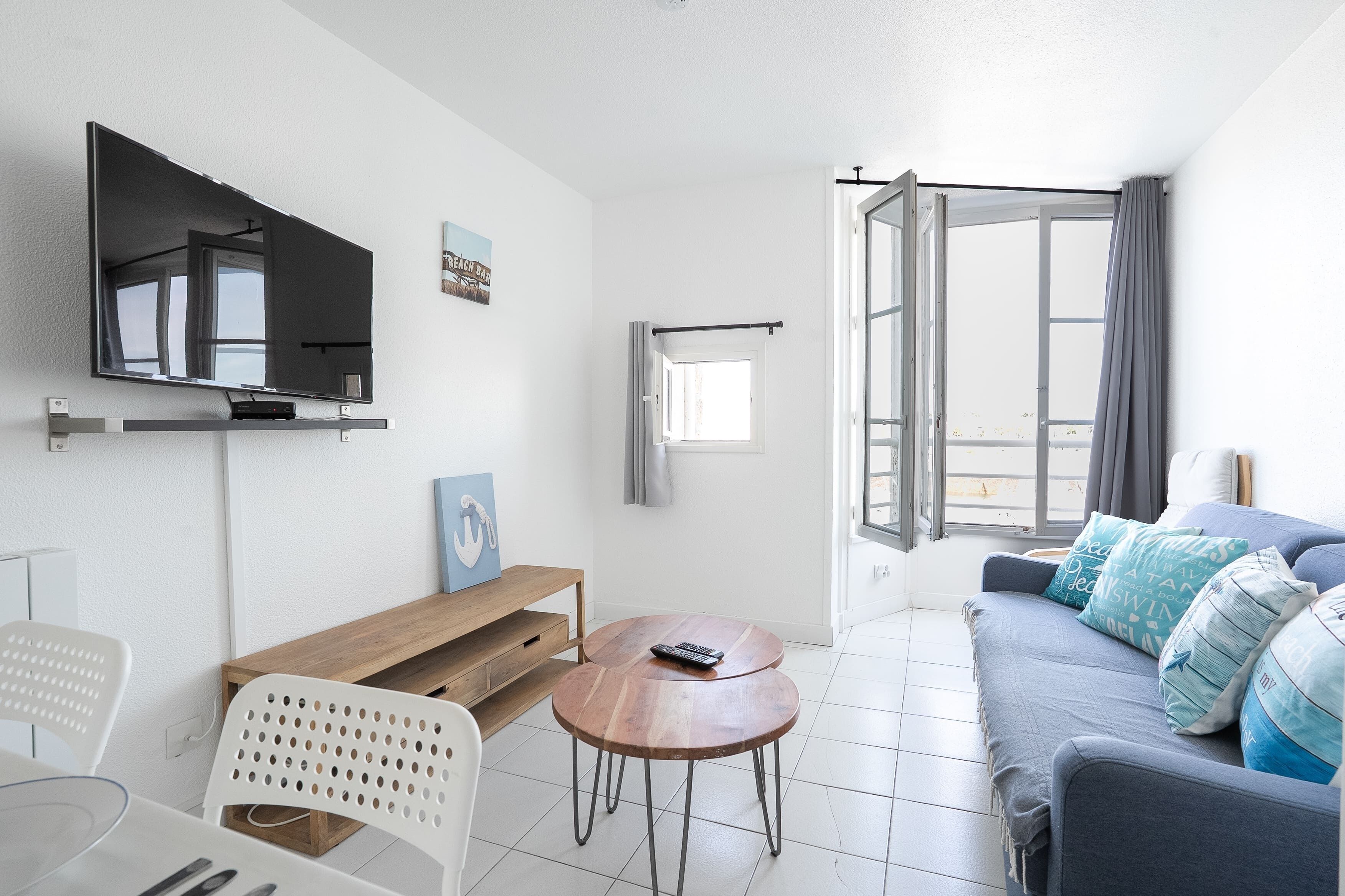 Ein modernes Wohnzimmer mit Flachbildfernseher, Couchtisch aus Holz, grauem Sofa und einem Essbereich mit weißem Stuhl und Tisch.