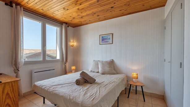 A bedroom with a wooden ceiling, a bed with a white bedspread, two bedside lamps, a radiator, and a window with curtains.
