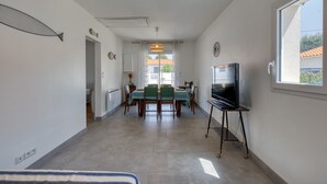 A spacious room with a dining area, a television on a stand, and a large window providing natural light.