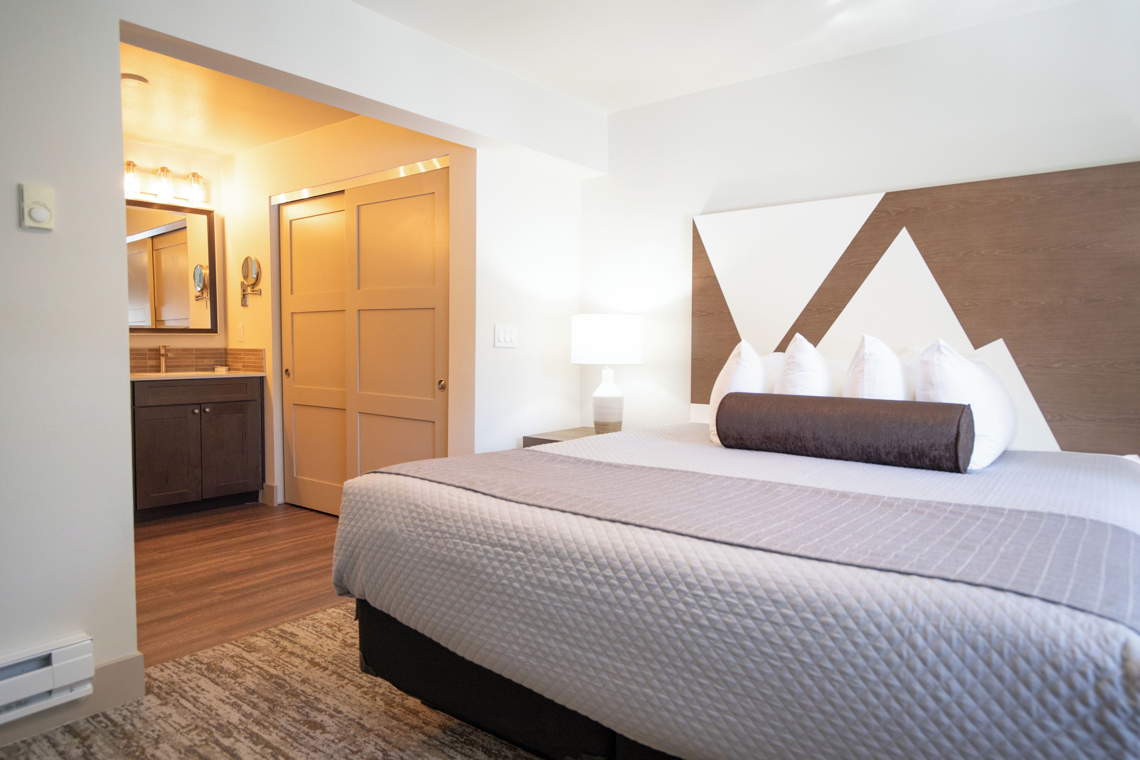 Quarto de hotel com uma cama, mesa de cabeceira, abajur, cômoda e banheiro visível por uma porta aberta.