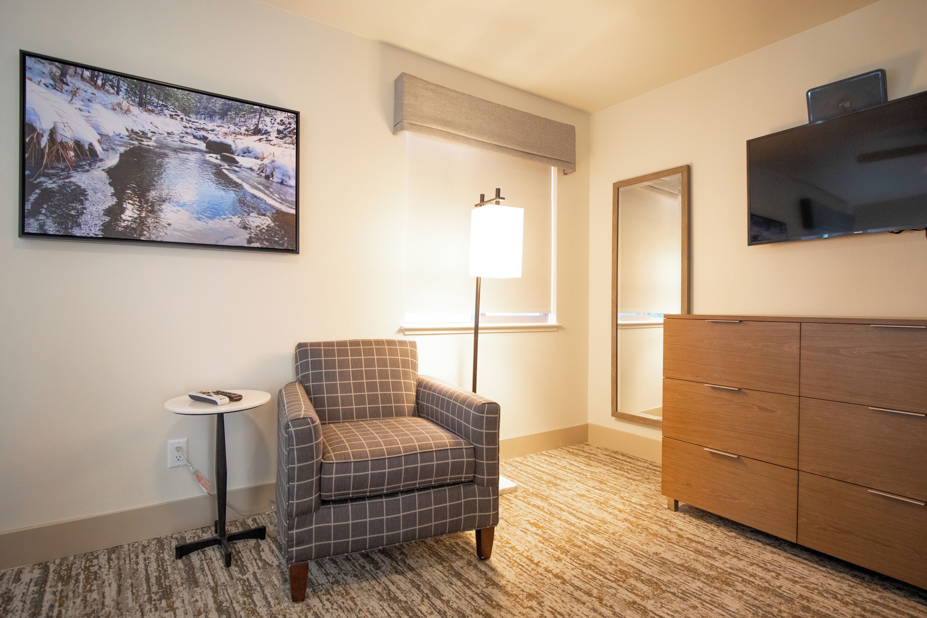 A hotel room with a plaid armchair, a small table with a remote control, a dresser, a large mirror, and a painting of a snowy landscape.