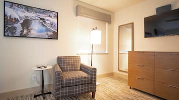 A hotel room with a plaid armchair, a small table with a remote control, a dresser, a large mirror, and a painting of a snowy landscape.