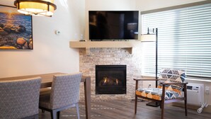 A living room with a fireplace, a flat-screen TV mounted above it, a dining table with chairs, and a large window with blinds. - Tahoe Trail (Stateline)