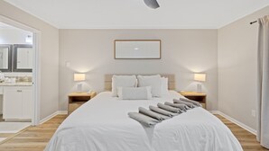 A neatly arranged bedroom with a large bed, two bedside tables, a framed artwork above the bed, and a view of the bathroom and kitchen through an open door.