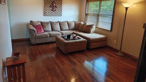 A living room with a sectional sofa, a coffee table, wooden flooring, a ceiling fan, a window with blinds, and a floor lamp. - Pointer Palace (South Point)