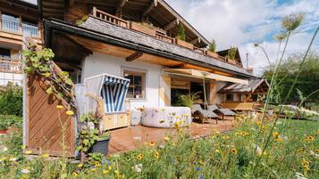 A traditional chalet with a wooden balcony, a covered patio with outdoor seating, and a garden with wildflowers.