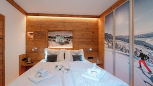 Quarto de hotel com cabeceira de madeira, uma grande moldura com uma imagem de paisagem nevada, cama com roupa de cama branca e um mural com tema de esqui na parede.